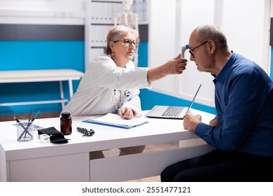 Old physician using thermometer on forehead of elderly man in hospital room, assessing his overall health. Retired female doctor checking temperature of pensioner patient during clinic appointment. - Powered by Shutterstock