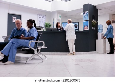 Old Physician Working With Asian Receptionist To Plan Checkup Report Papers At Hospital Reception Desk. Medic Taking To Clinic Worker At Counter About Medical Forms And Healthcare Insurance Support.