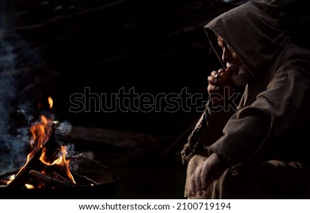 Old poor man sitting by the fire with a flute in the Middle Ages