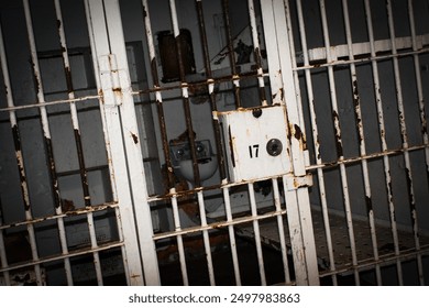 old prison bars showing a prison cell locked with bed and toilet. The bars are old and rusted. The cell door is locked with the number 17 painted on it. The cell is dark and gloomy.