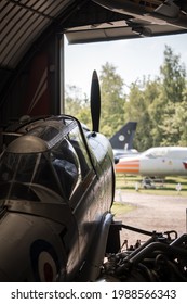 Old Propeller Aeroplane Fighter Plane Parked In Hanger On Sunny Day Looking Out Towards Airfield Runway And Other Jets. Dark Shadows And Canopy Reflections.