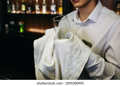 Old Pub With Bartender Cleaning The Glass With White Towel On Background. Empty Name Tag Badge On The Shirt Uniform. Barman At His Working Place.