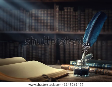 Old quill pen, books and vintage inkwell on wooden desk in the old office against the background of the bookcase and the rays of light. Conceptual background on history, education, literature topics.