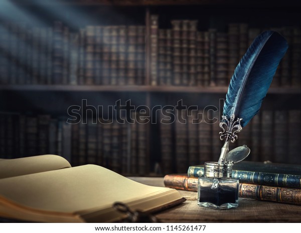 Old quill pen, books and vintage inkwell on\
wooden desk in the old office against the background of the\
bookcase and the rays of light. Conceptual background on history,\
education, literature\
topics.