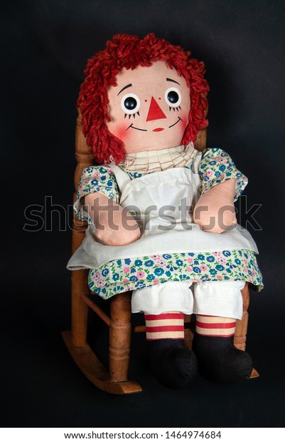 old rag doll with white apron sitting in a wooden\
rocking chair