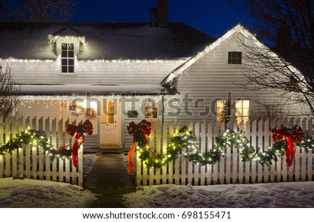 An old ranch house, complete with white picket fence, is decked out in Christmas lights, ribbons and garlands.  A Christmas tree in the window sets the scene for a romantic country Christmas.