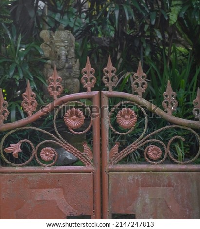 Old red iron gate with flower emblems and blurred background with Balinese ganesha concrete statue among green plants
