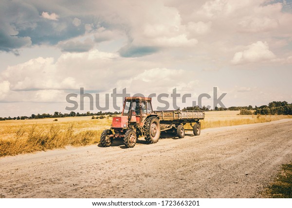 Old Red\
Tractor with a Trailer moving on the Countryside Sandy Road in hot\
summer day. Father and Son inside a tractor.\
