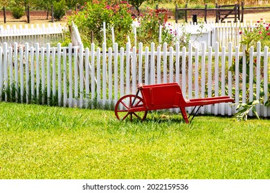 Old red wooden wheel barrel in a garden with a white picket fence #1