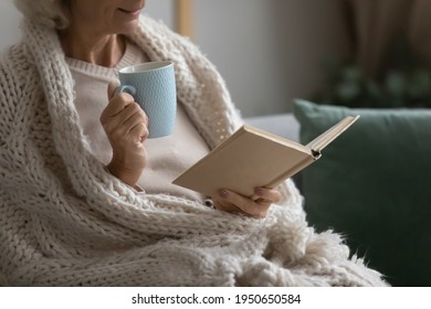 Old retired 70s lady reading book at home, drinking hot tea, resting on sofa in cozy living room. Mature reader wrapped in knitted blanket relaxing with novel over cup of coffee or chocolate. Close up
