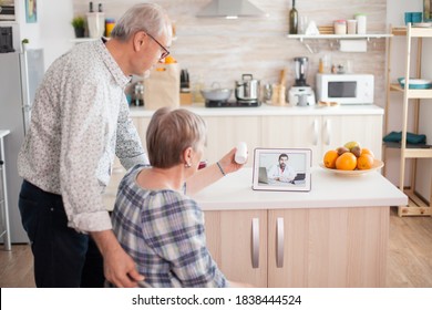 Old Retired Couple Checking The Prescription With Doctor Via Online Video Call. Video Conference With Doctor Using Laptop In Kitchen. Online Health Consultation For Elderly People Drugs Ilness Advice