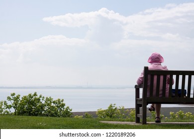 An Old Retired English Woman In Pink Clothes Sitting On The Bench And Enjoying Seascape View.