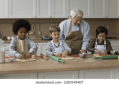 Old retired grandfather teaching diverse grandkids to bake in home kitchen, rolling, cutting, shaping dough on floury table, preparing homemade dessert for family dinner - Powered by Shutterstock