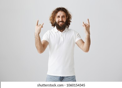 Old Rock Still Lives In My Soul. Excited And Emotive Easter Man With Long Curly Hair And Beard Showing Rock Gestures While Being On Concert Of His Favourite Group, Enjoying Great Music Over Gray Wall