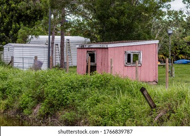 Old Rotting Faded Painted Pink Shingle Roof Barn Style Small Shed With White Trim Open Door And Window On A Backyard Farm Next To Metal Storage, A-frame Ladders In Tall Weeds & Grass Found In Florida