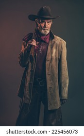 Old Rough Western Cowboy With Gray Beard And Brown Hat Holding Rifle. Low Key Studio Shot.