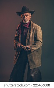 Old Rough Western Cowboy With Gray Beard And Brown Hat Holding Rifle. Low Key Studio Shot.