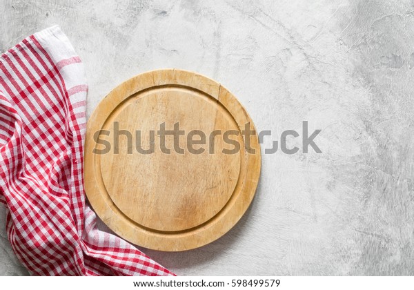 Old round wooden cutting board (pizza board) over\
bright gray stone background and classic red checkered tablecloth.\
Copy space for text. Cooking food, pizza, background. Design mock\
up.