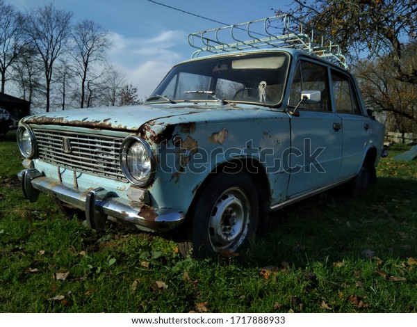 An old, ruined,\
damaged soviet car on the background of the rural abandoned\
landscape. Blue summer sky