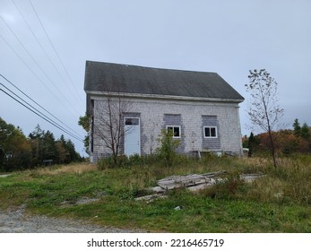 An old rundown and unused building sitting on a overgrown lawn. Random boards are plastered up over the building and the roof shingles are weathered.