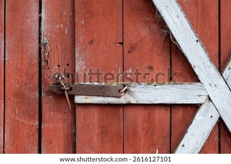 Old rustic latch with a chain securing a weathered red shed door, framed by faded white wooden beams and covered in cobwebs, showing age and rural charm