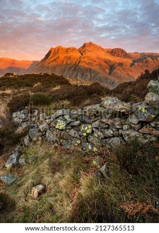 Old rustic stone wall standing against the harsh mountainous environment high up in the Lake District mountains. The Langdale Pikes in the background, catching golden light from morning sun.