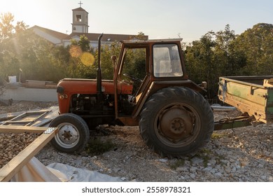 Old rustic tractor with trailer parked on rocky ground under golden evening sunlight near rural church and greenery. Concept of countryside life, farming equipment, and nostalgic agriculture scenes.  - Powered by Shutterstock