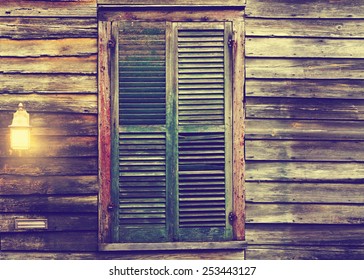 Old Rustic Vintage Antique Dilapidated Run Down Leaning House Home Building Structure With Green Closed Shut Shutters Over Window And Porch Light Lantern Glowing Turned On 