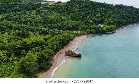 An old, rusting shipwreck stranded on a sandy beach surrounded by a dense green tropical forest
