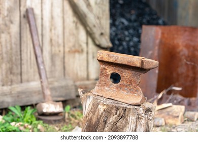 Old Rusty Anvil Stands On A Stump In The Backyard Of Village House, Sledgehammer Leaning On Wooden Fence Enclosing A Barn With Coal On Blurred Background. Heavy Working Tools.