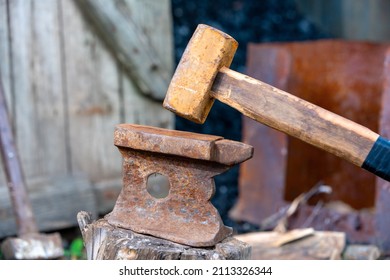 Old Rusty Anvil Stands On A Stump In The Backyard Of Village House, Sledgehammer Leaning On Wooden Fence Enclosing A Barn With Coal On Blurred Background. Heavy Working Tools