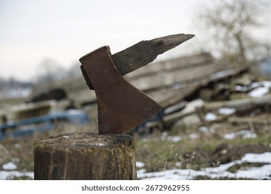 An old rusty axe is stuck in a tree stump on a snowy day, with rubble piles in the background. The axe handle is also damaged.