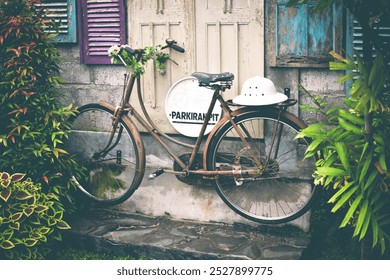 An old, rusty bicycle leans against a wall with colorful, weathered shutters. A white helmet rests on its seat, and green foliage wraps around the handlebars. - Powered by Shutterstock