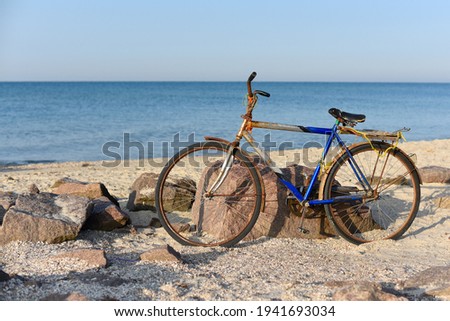An old rusty bicycle is parked near the rocks on the seashore. The blurry background is the blue calm sea. Recreation. Sports. Fishing. Hobby. Sunny day. 