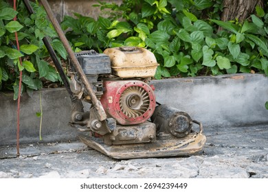 Old and rusty construction compactor machine left on broken concrete path near green plants in outdoor setting
