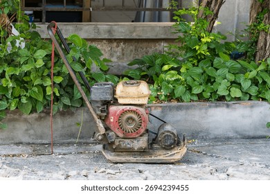 Old and rusty construction compactor machine left on broken concrete path near green plants in outdoor setting