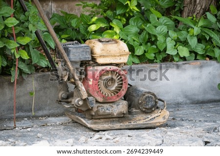 Old and rusty construction compactor machine left on broken concrete path near green plants in outdoor setting