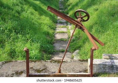 Old Rusty Crank In An Abandoned Dam. View Of The Dry Canal, Water Shortage, In The Green Meadow
