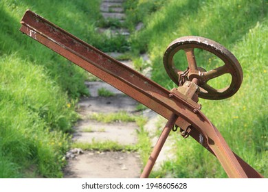 Old Rusty Crank In An Abandoned Dam. View Of The Dry Canal, Water Shortage, In The Green Meadow
