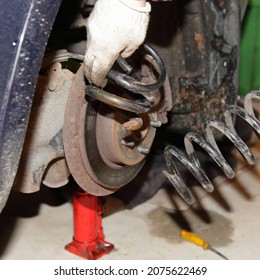 Old Rusty Damaged Coil Spring In Hands On Broken Car On Bottle Hydraulic Jack Background - Vehicle Suspension Repair In Garage