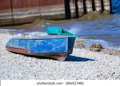 Old Rusty Fishing Boat Lies On The Pebble Coast Of The River. The Waves Of The Tide Roll Over The Little Iron Man And Break Into Spray And Foam On The Side, Bow And Stern Of The Boat.