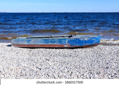 Old Rusty Fishing Boat Lies On The Pebble Coast Of The River. The Waves Of The Tide Roll Over The Little Iron Man And Break Into Spray And Foam On The Side, Bow And Stern Of The Boat.