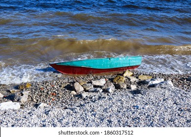 Old Rusty Fishing Boat Lies On The Pebble Coast Of The River. The Waves Of The Tide Roll Over The Little Iron Man And Break Into Spray And Foam On The Side, Bow And Stern Of The Boat.