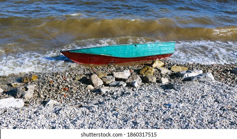 Old Rusty Fishing Boat Lies On The Pebble Coast Of The River. The Waves Of The Tide Roll Over The Little Iron Man And Break Into Spray And Foam On The Side, Bow And Stern Of The Boat.