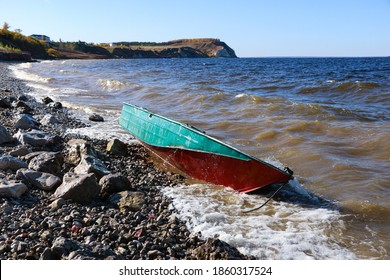 Old Rusty Fishing Boat Lies On The Pebble Coast Of The River. The Waves Of The Tide Roll Over The Little Iron Man And Break Into Spray And Foam On The Side, Bow And Stern Of The Boat.