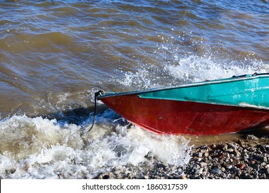 Old Rusty Fishing Boat Lies On The Pebble Coast Of The River. The Waves Of The Tide Roll Over The Little Iron Man And Break Into Spray And Foam On The Side, Bow And Stern Of The Boat.