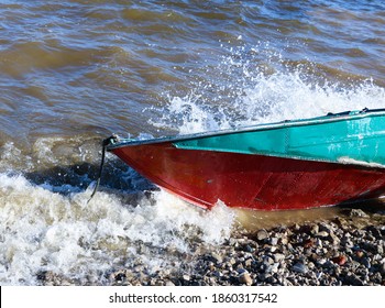 Old Rusty Fishing Boat Lies On The Pebble Coast Of The River. The Waves Of The Tide Roll Over The Little Iron Man And Break Into Spray And Foam On The Side, Bow And Stern Of The Boat.