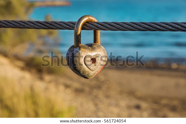 Old\
rusty heart shaped padlock with peeled off red paint in enlarged\
view as symbol of eternal love hanging on bridge metal cable in\
front of beach. Romance concept. Blurred background.\
