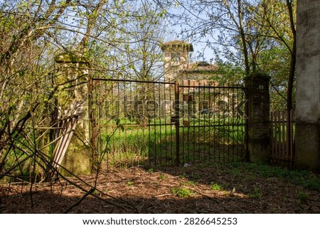 An old rusty iron gate is closed between two stone pillars covered in moss. In the background, beyond the overgrown garden and wild trees, a large abandoned vintage villa with a panoramic tower structure can be glimpsed.