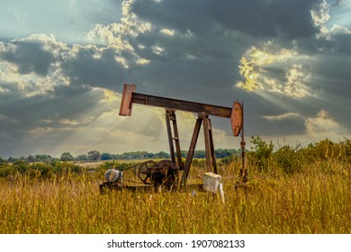 Old Rusty Oil Well Pump Jack In Field - Home Made Modifications - With Field And Cows Blurred In Background And Dramatic Stormy Evening Sky